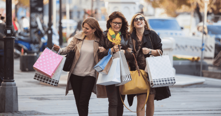 3 Women with linked arms walking outdoors carrying handbags and shopping bags.