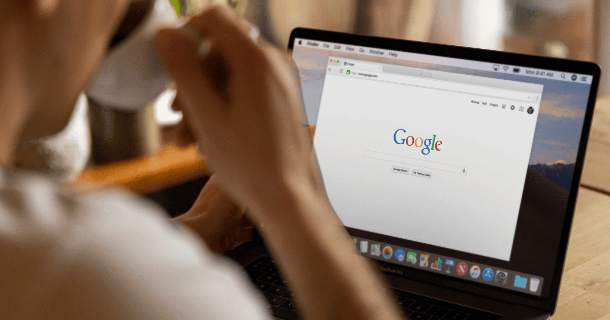 Person seated at table drinking from a cup with laptop screen open viewing a Google search page. 